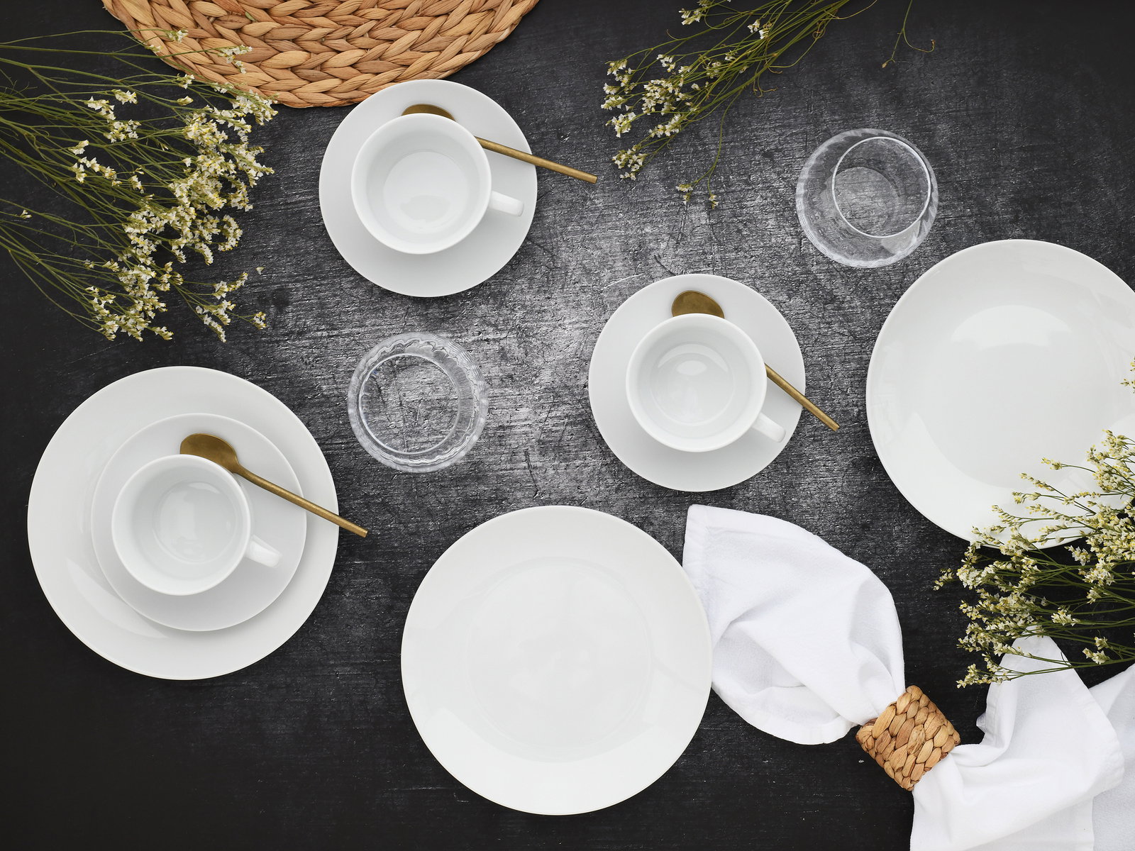 Vogelperspektive auf ein elegantes, weißes 18-teiliges Kaffeeservice mit goldenen Löffeln, umgeben von zarten weißen Blumen und einem geflochtenen Tischset.