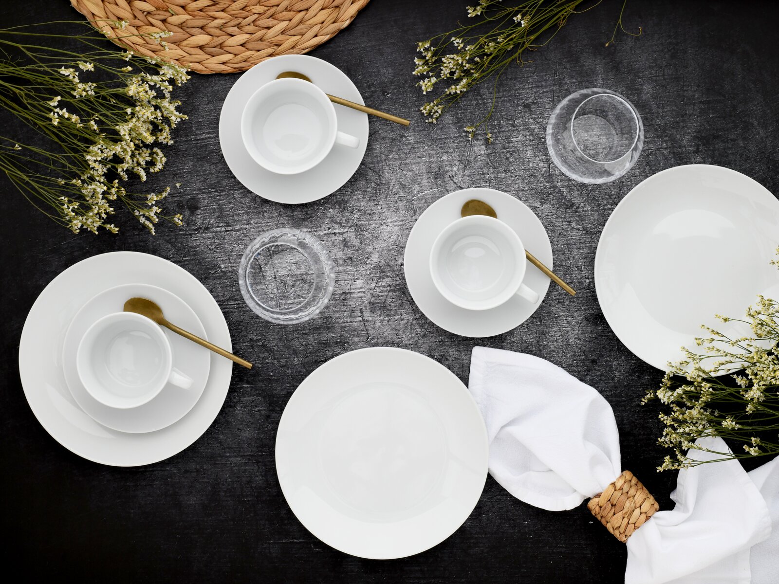 Vogelperspektive auf ein elegantes, weißes 18-teiliges Kaffeeservice mit goldenen Löffeln, umgeben von zarten weißen Blumen und einem geflochtenen Tischset.