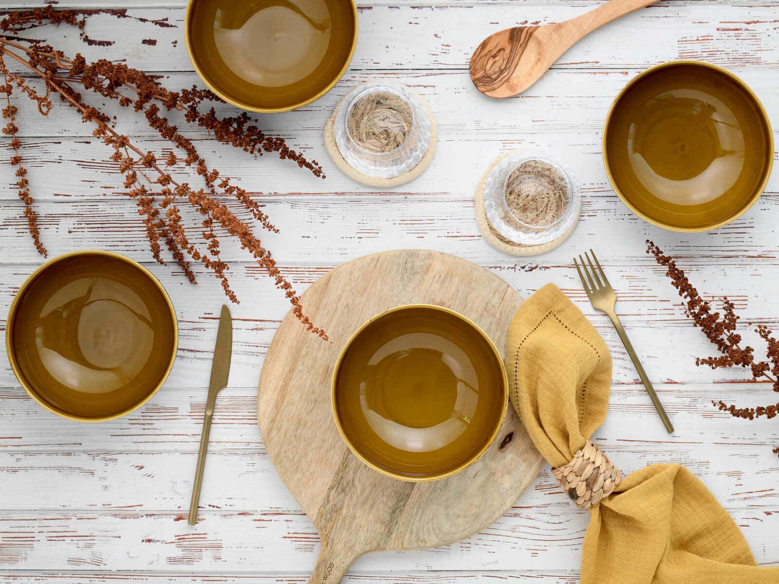 Draufsicht auf ein stilvolles Tisch-Arrangement mit mehreren olivgrünen Keramikschalen, Holzbrett, Holzlöffel, goldenem Besteck, gelber Stoffserviette und dekorativen Trockenblumen auf weißer Holzoberfläche.