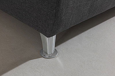 Nahaufnahme eines Boxspringbett-Fußes aus Metall auf einem hellen Teppichboden, perspektivisch von der Seite aufgenommen.