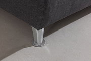 Nahaufnahme eines Boxspringbett-Fußes aus Metall auf einem hellen Teppichboden, perspektivisch von der Seite aufgenommen.