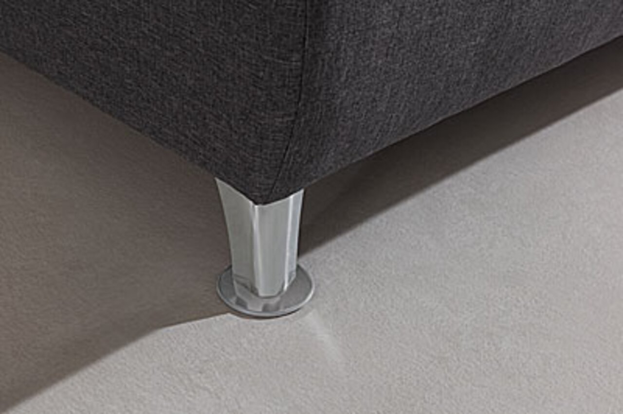 Nahaufnahme eines Boxspringbett-Fußes aus Metall auf einem hellen Teppichboden, perspektivisch von der Seite aufgenommen.