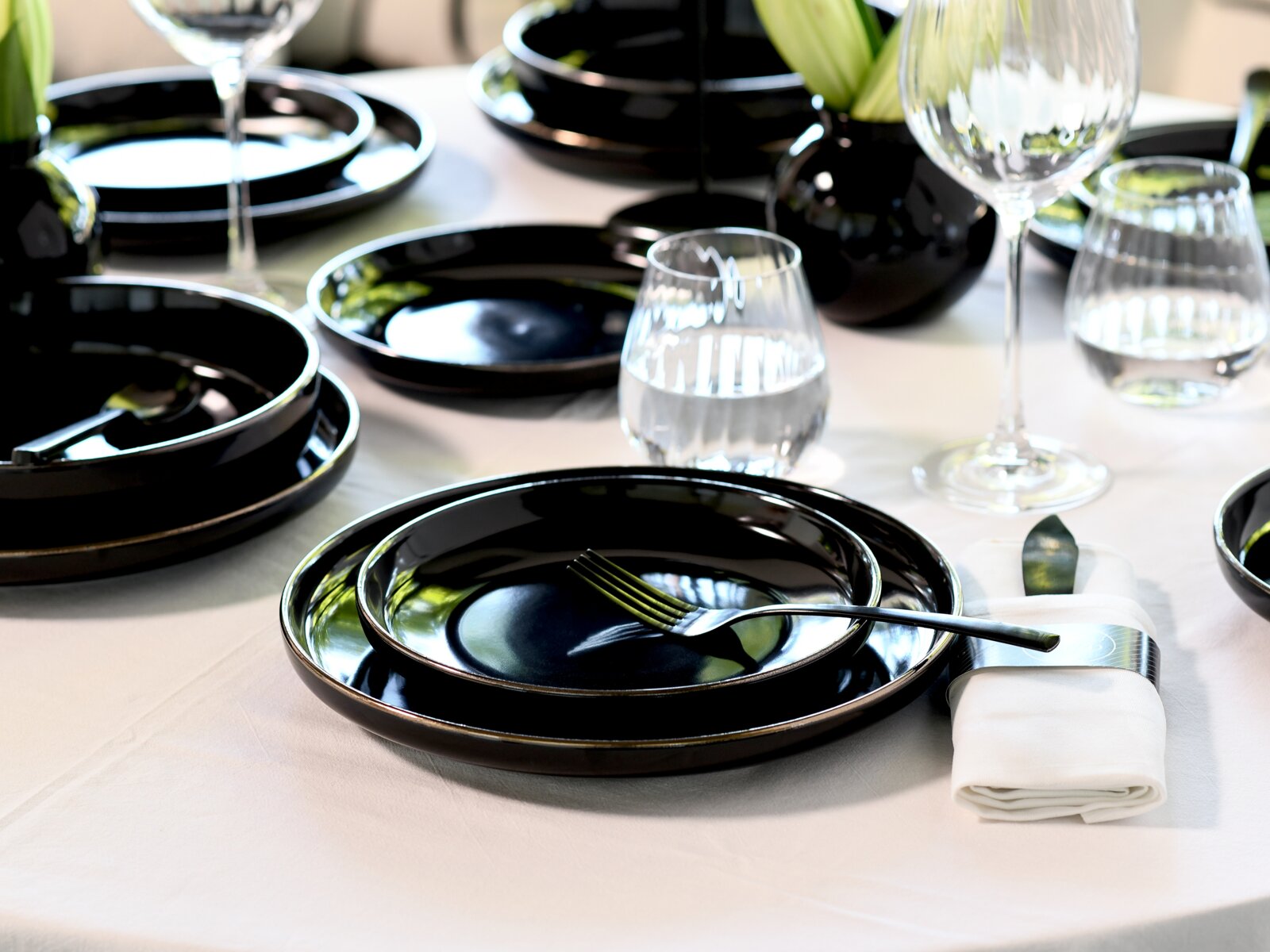 Elegant gedeckter Tisch mit glänzend schwarzen Tellern und Schalen, Besteck, gefalteter weißer Serviette sowie Wasser- und Weingläsern; Perspektive: leicht schräg von oben in Nahaufnahme.