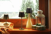Drei Akku-Tischleuchten VANNIE 3 in verschiedenen Größen auf einer Fensterbank, umgeben von einer Laterne und einem Geschenk, mit Blick nach draußen.
