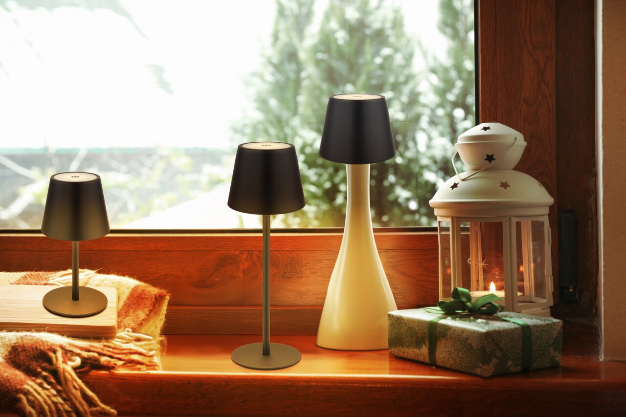 Drei Akku-Tischleuchten VANNIE 3 in verschiedenen Größen auf einer Fensterbank, umgeben von einer Laterne und einem Geschenk, mit Blick nach draußen.