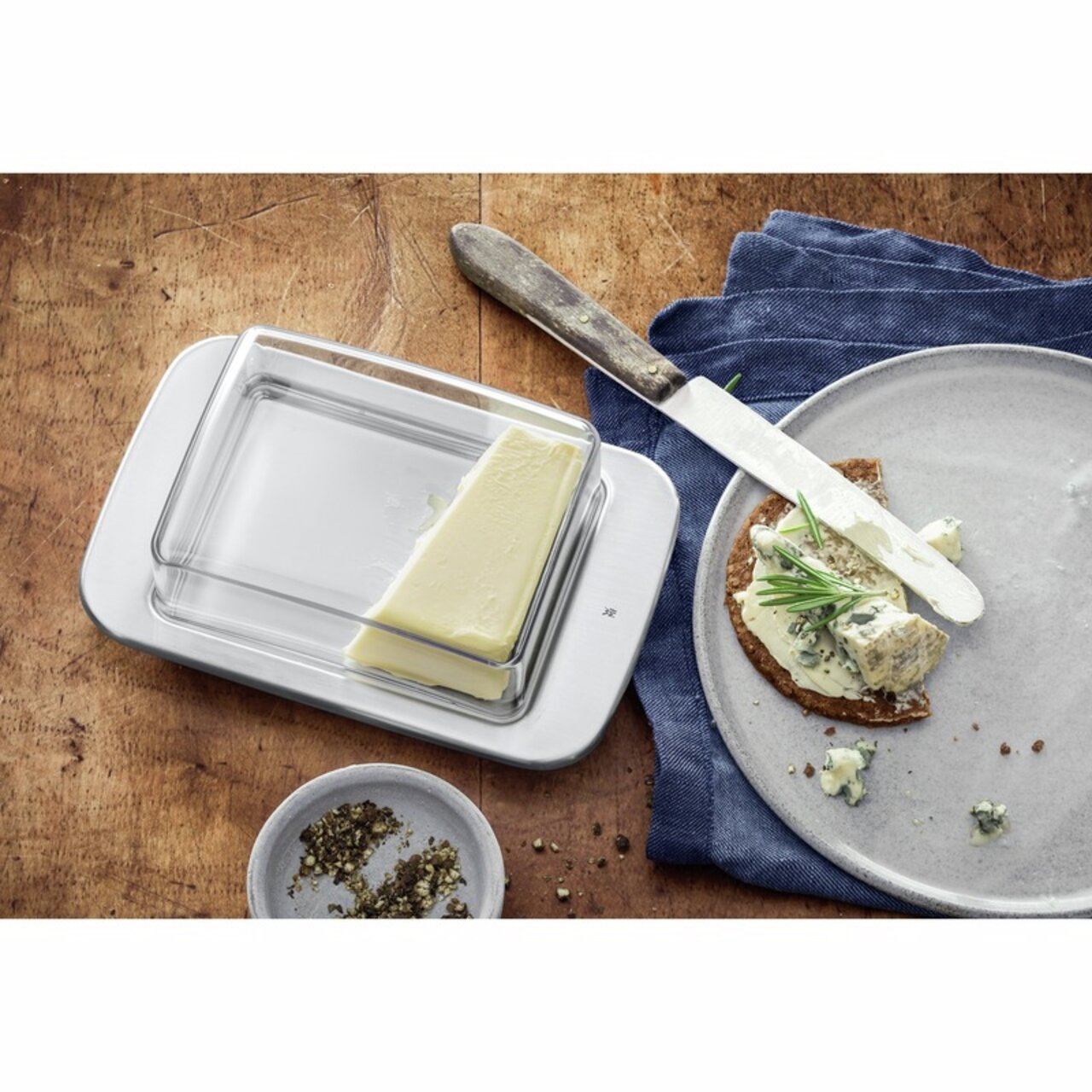 WMF Butterdose LOFT BRUNCH Butterdose LOFT aus Glas und Metall, von oben fotografiert, mit einem Stück Butter auf einem Holztisch neben einem Teller mit Brot und Käse.