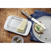 Butterdose LOFT aus Glas und Metall, von oben fotografiert, mit einem Stück Butter auf einem Holztisch neben einem Teller mit Brot und Käse.