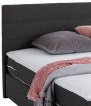 Nahaufnahme eines Boxspringbetts mit grauem Kopfteil, grauem Kissen und rosa Decke, seitliche Perspektive