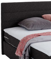 Nahaufnahme eines Boxspringbetts mit grauem Kopfteil, grauem Kissen und rosa Decke, seitliche Perspektive