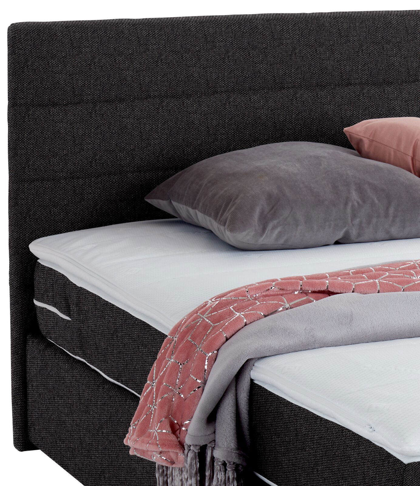 Nahaufnahme eines Boxspringbetts mit grauem Kopfteil, grauem Kissen und rosa Decke, seitliche Perspektive