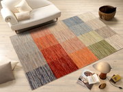 Bunte Wollteppich mit quadratischem Muster in verschiedenen Farben, auf einem hellen Holzboden neben einem weißen Sofa und einem Korb, aus der Vogelperspektive betrachtet.