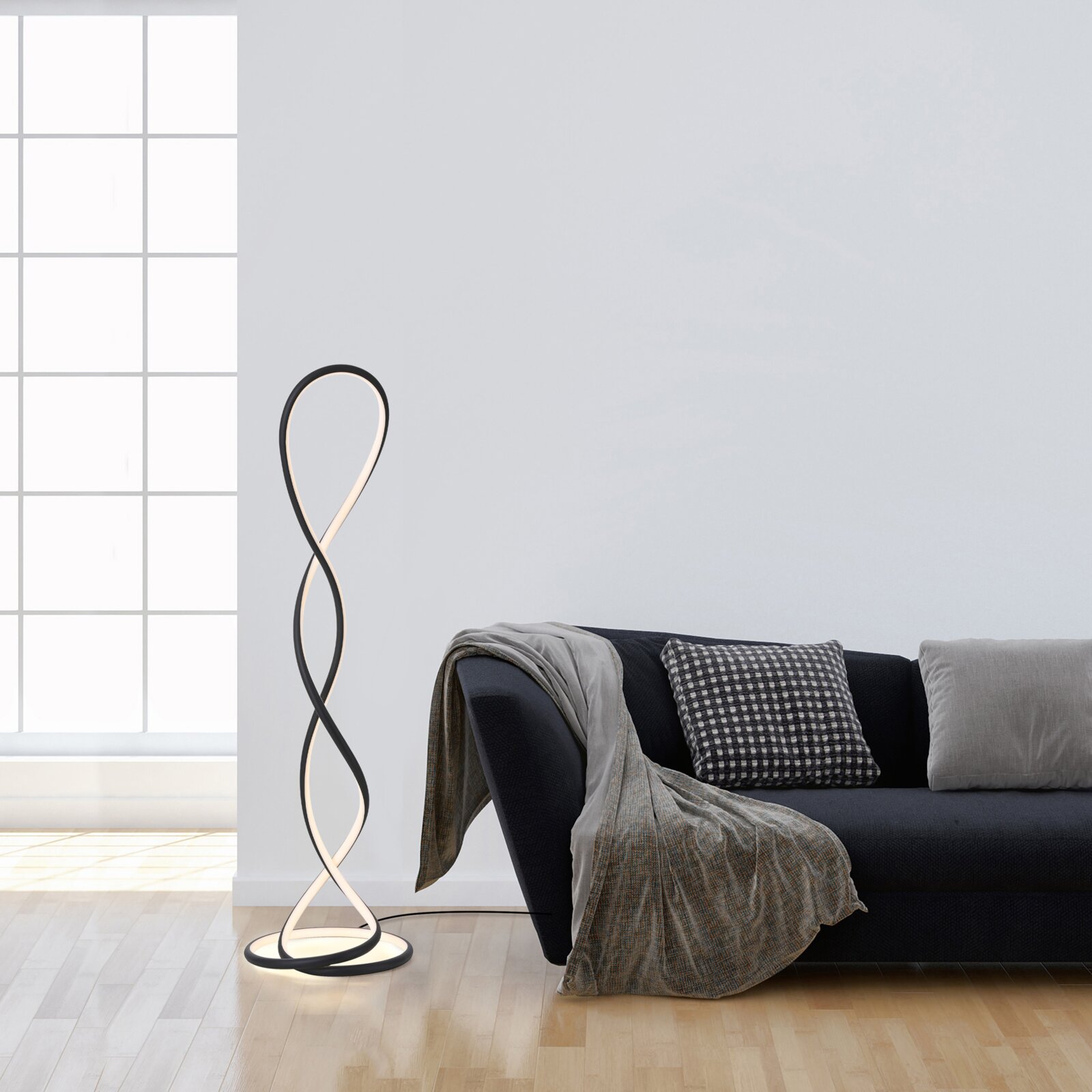 Moderne LED-Stehleuchte in schwarzem Metall mit geschwungenem, doppelt gedrehtem Lichtband und rundem Sockel neben einem dunklen Sofa im Wohnzimmer, perspektivisch in leichter Seitenansicht auf Augenhöhe