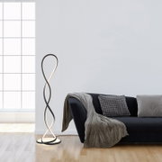 Moderne LED-Stehleuchte in schwarzem Metall mit geschwungenem, doppelt gedrehtem Lichtband und rundem Sockel neben einem dunklen Sofa im Wohnzimmer, perspektivisch in leichter Seitenansicht auf Augenhöhe