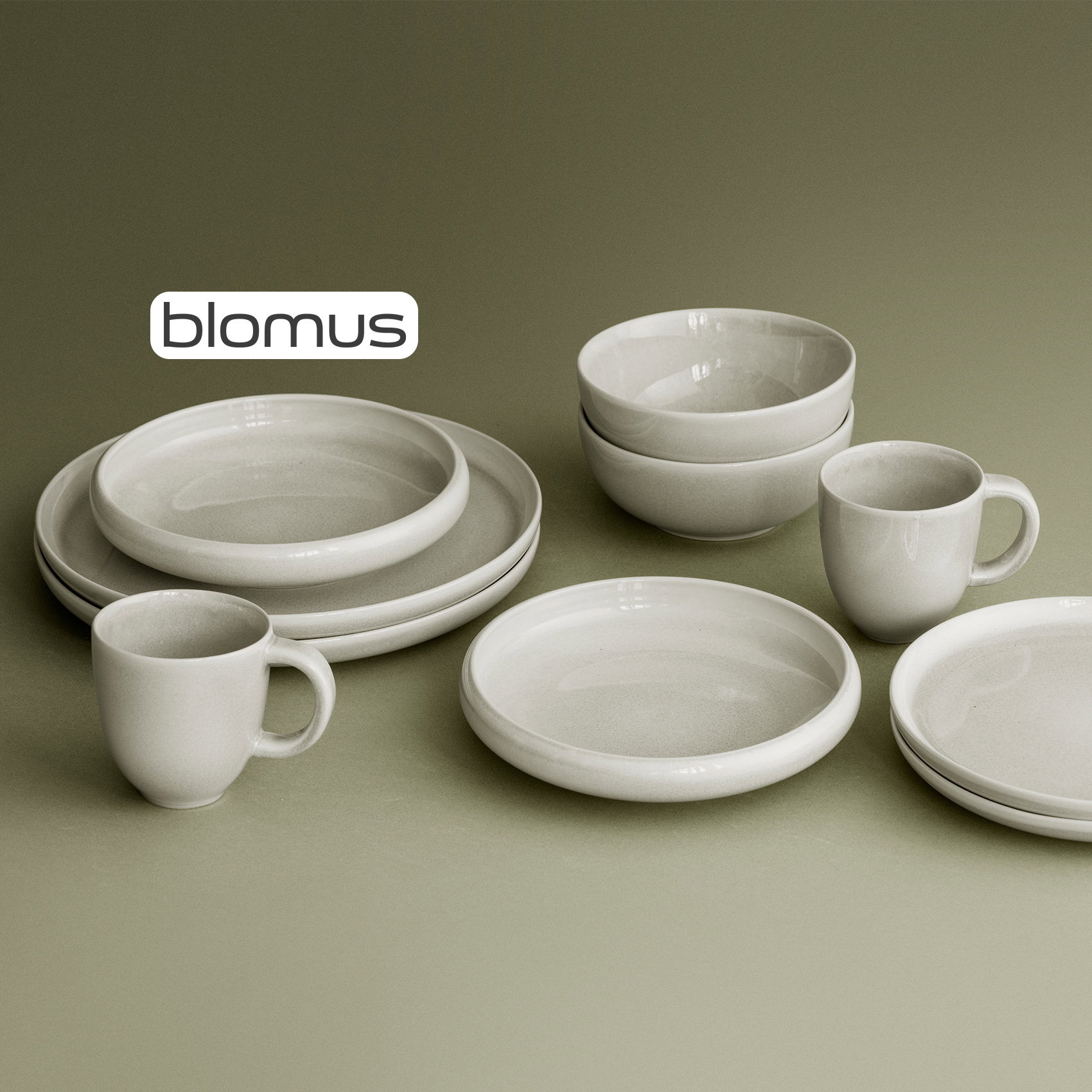Helles Blomus-Geschirr mit Tellern, Schalen und Tassen in natürlicher Keramikoptik auf olivgrünem Hintergrund.