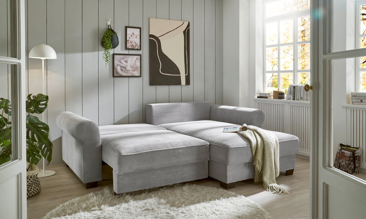 Gemütliche graue Eckcouch in einem hellen Wohnzimmer mit moderner Dekoration, aus der Perspektive von vorne links betrachtet.