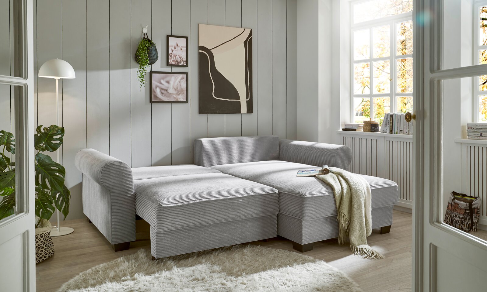 Gemütliche graue Eckcouch in einem hellen Wohnzimmer mit moderner Dekoration, aus der Perspektive von vorne links betrachtet.