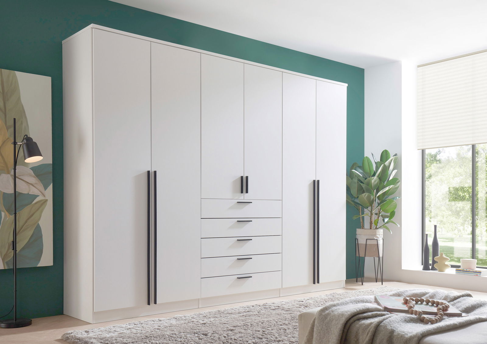 Moderner, weißer 6-türiger Drehtürenschrank mit schwarzen Griffen, frontal fotografiert in einem stilvoll eingerichteten Raum mit grüner Wand und großen Fenstern.
