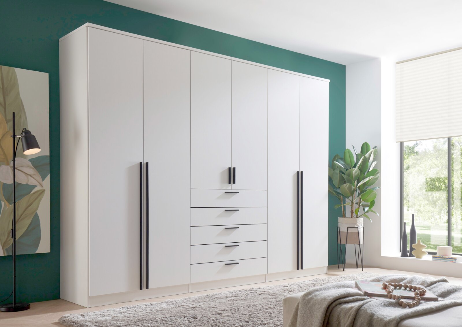 Moderner, weißer 6-türiger Drehtürenschrank mit schwarzen Griffen, frontal fotografiert in einem stilvoll eingerichteten Raum mit grüner Wand und großen Fenstern.