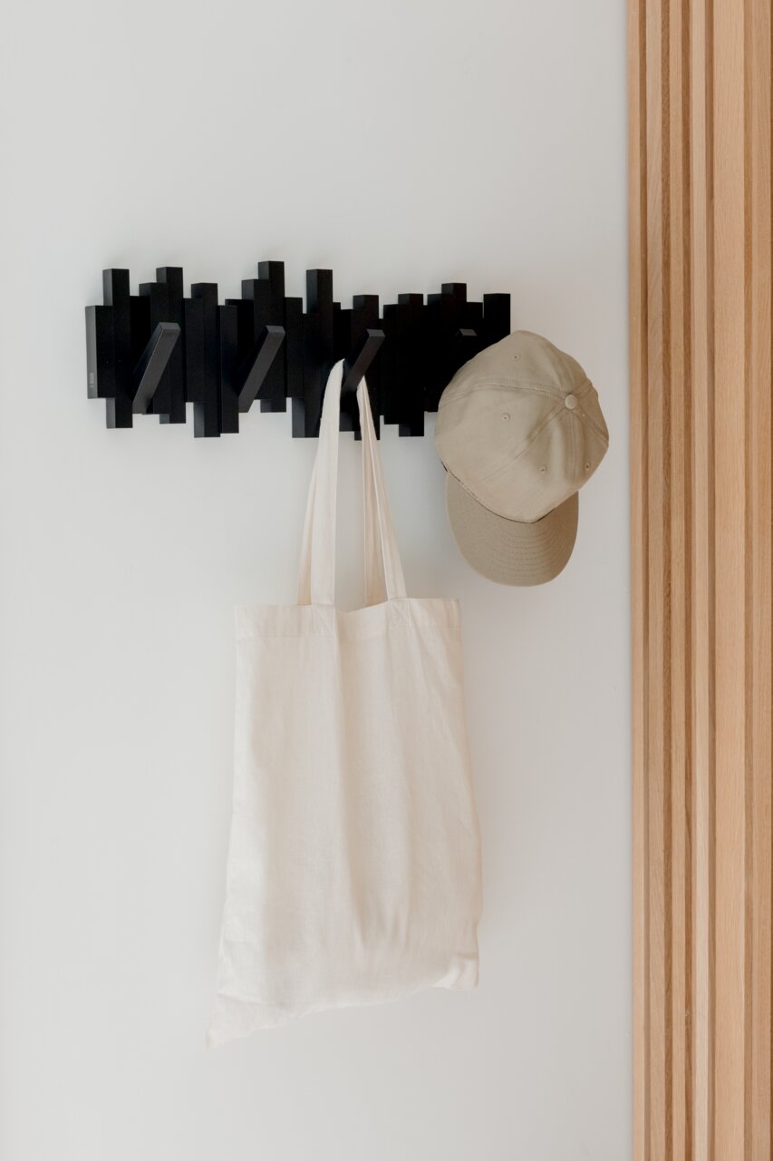 Schwarze Wandgarderobe mit mehreren Haken, an der eine beige Kappe und eine weiße Stofftasche hängen, seitliche Perspektive.
