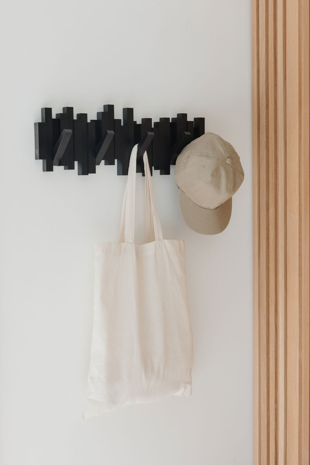 Schwarze Wandgarderobe mit mehreren Haken, an der eine beige Kappe und eine weiße Stofftasche hängen, seitliche Perspektive.