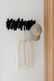 Schwarze Wandgarderobe mit mehreren Haken, an der eine beige Kappe und eine weiße Stofftasche hängen, seitliche Perspektive.