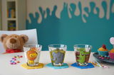 Drei bunte Kindertrinkgläser mit Tiermotiven (Löwe, Elefant, Krokodil) auf einem Tisch, umgeben von Süßigkeiten und einem Plüschtier im Hintergrund. Frontalansicht.