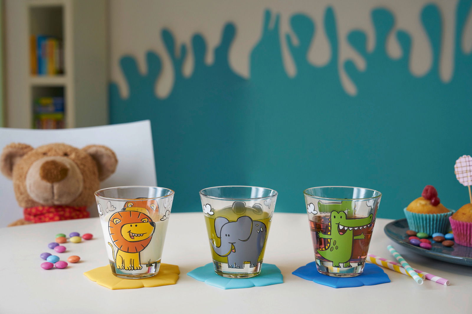Drei bunte Kindertrinkgläser mit Tiermotiven (Löwe, Elefant, Krokodil) auf einem Tisch, umgeben von Süßigkeiten und einem Plüschtier im Hintergrund. Frontalansicht.