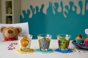 Drei bunte Kindertrinkgläser mit Tiermotiven (Löwe, Elefant, Krokodil) auf einem Tisch, umgeben von Süßigkeiten und einem Plüschtier im Hintergrund. Frontalansicht.