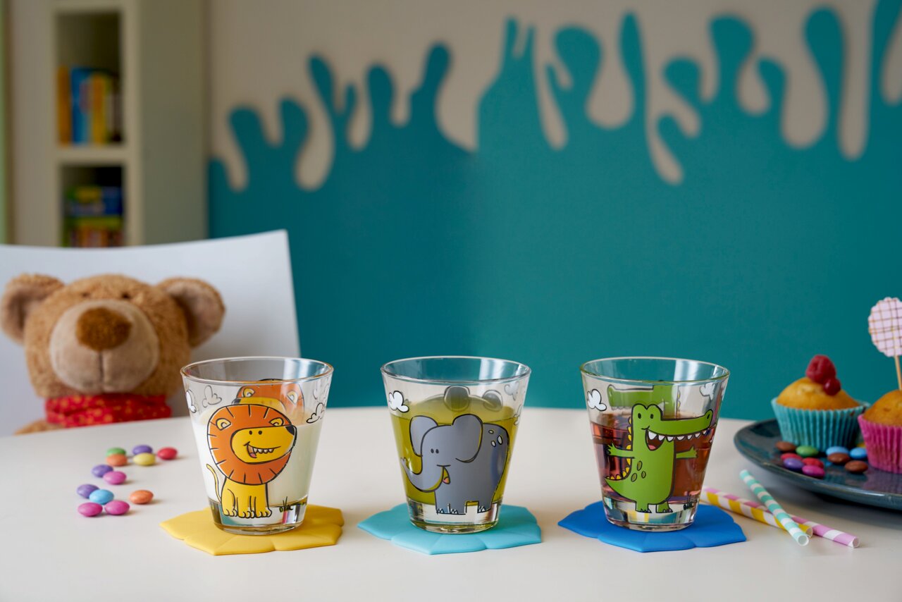 Drei bunte Kindertrinkgläser mit Tiermotiven (Löwe, Elefant, Krokodil) auf einem Tisch, umgeben von Süßigkeiten und einem Plüschtier im Hintergrund. Frontalansicht.