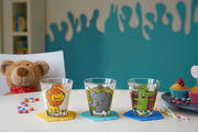 Drei bunte Kindertrinkgläser mit Tiermotiven (Löwe, Elefant, Krokodil) auf einem Tisch, umgeben von Süßigkeiten und einem Plüschtier im Hintergrund. Frontalansicht.