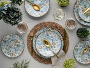 Draufsicht auf ein gedecktes Tischarrangement mit gemusterten Keramiktellern im Mosaikdesign in Blau- und Ockertönen, goldfarbenem Besteck, Glas und geflochtenem Platzset auf Leinentischdecke