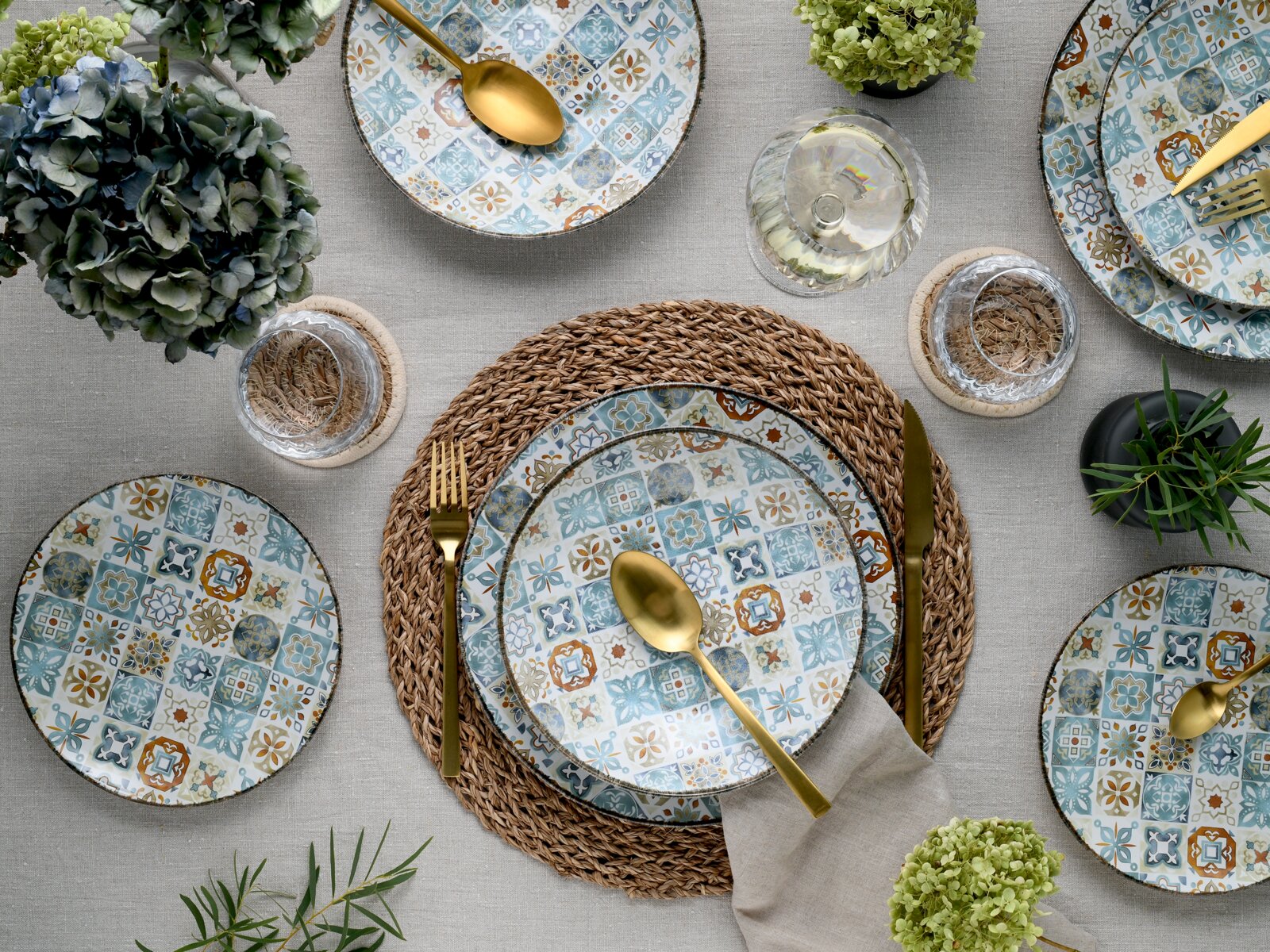 Draufsicht auf ein gedecktes Tischarrangement mit gemusterten Keramiktellern im Mosaikdesign in Blau- und Ockertönen, goldfarbenem Besteck, Glas und geflochtenem Platzset auf Leinentischdecke
