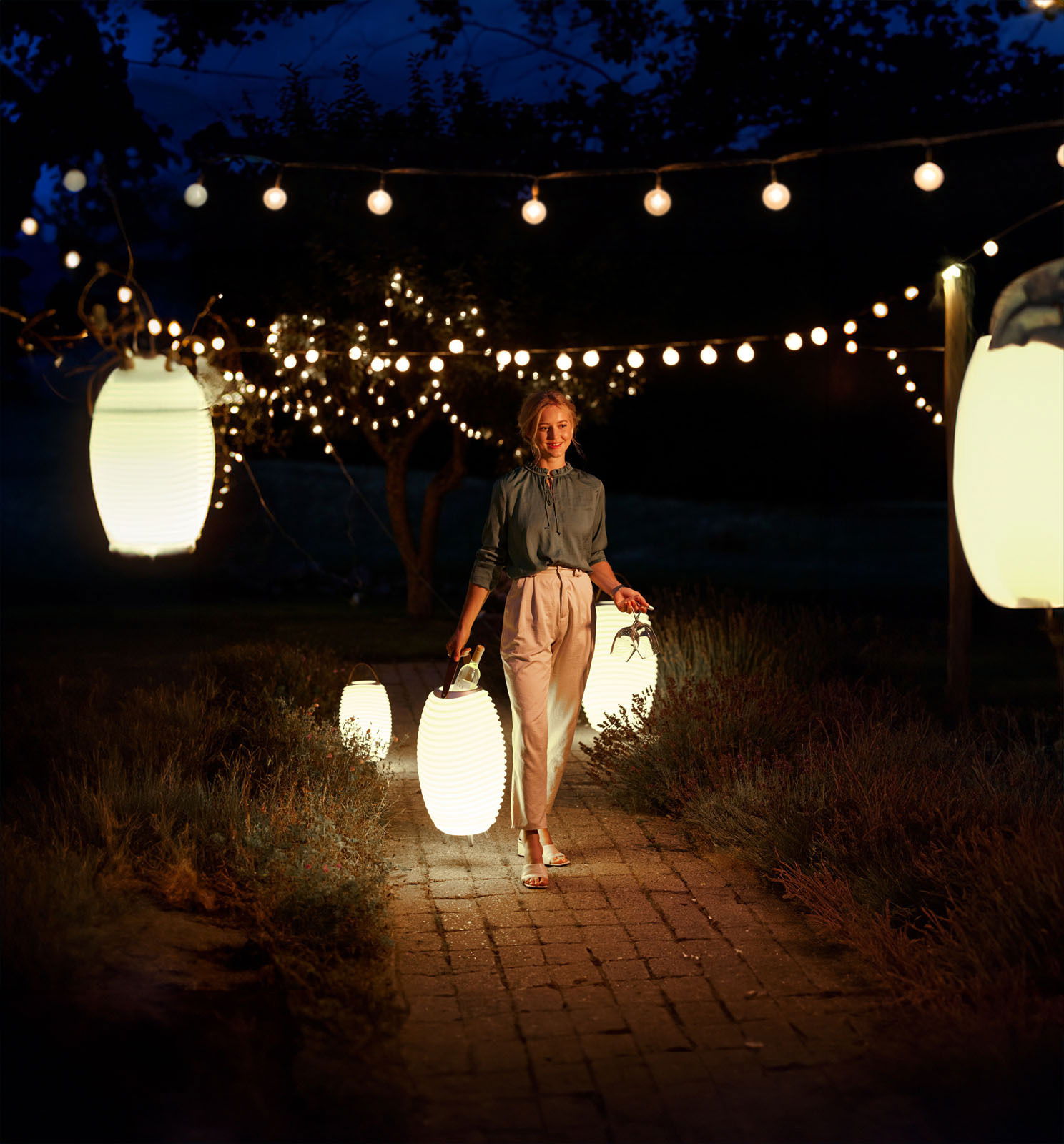 Frau trägt eine leuchtende Akku-Laterne in einem stimmungsvoll beleuchteten Garten bei Nacht, umgeben von hängenden Lichterketten.