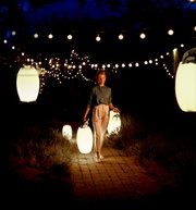 Een vrouw draagt een felle acculantaarn in een sfeervol verlichte tuin 's nachts, omringd door hangende lichtjes.