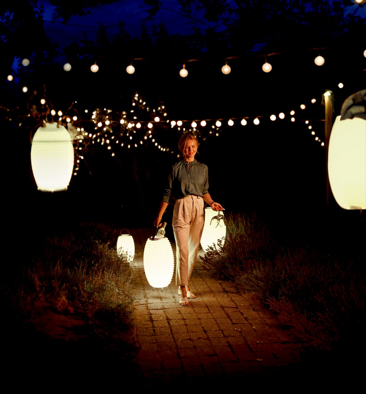 Een vrouw draagt een felle acculantaarn in een sfeervol verlichte tuin 's nachts, omringd door hangende lichtjes.