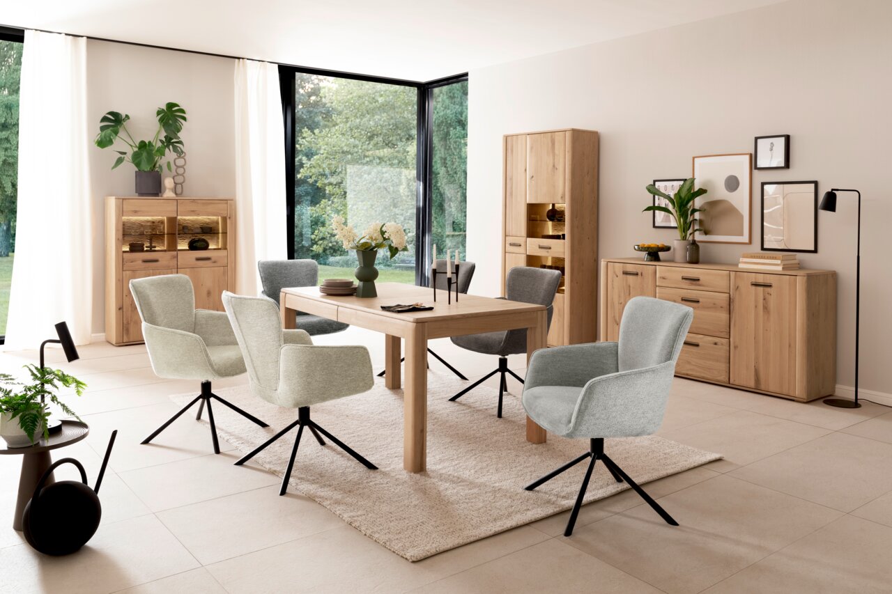 Moderne Esszimmer-Einrichtung mit Holztisch, gepolsterten Drehstühlen und Vitrinen in einem hellen Raum mit großen Fenstern, seitliche Perspektive.