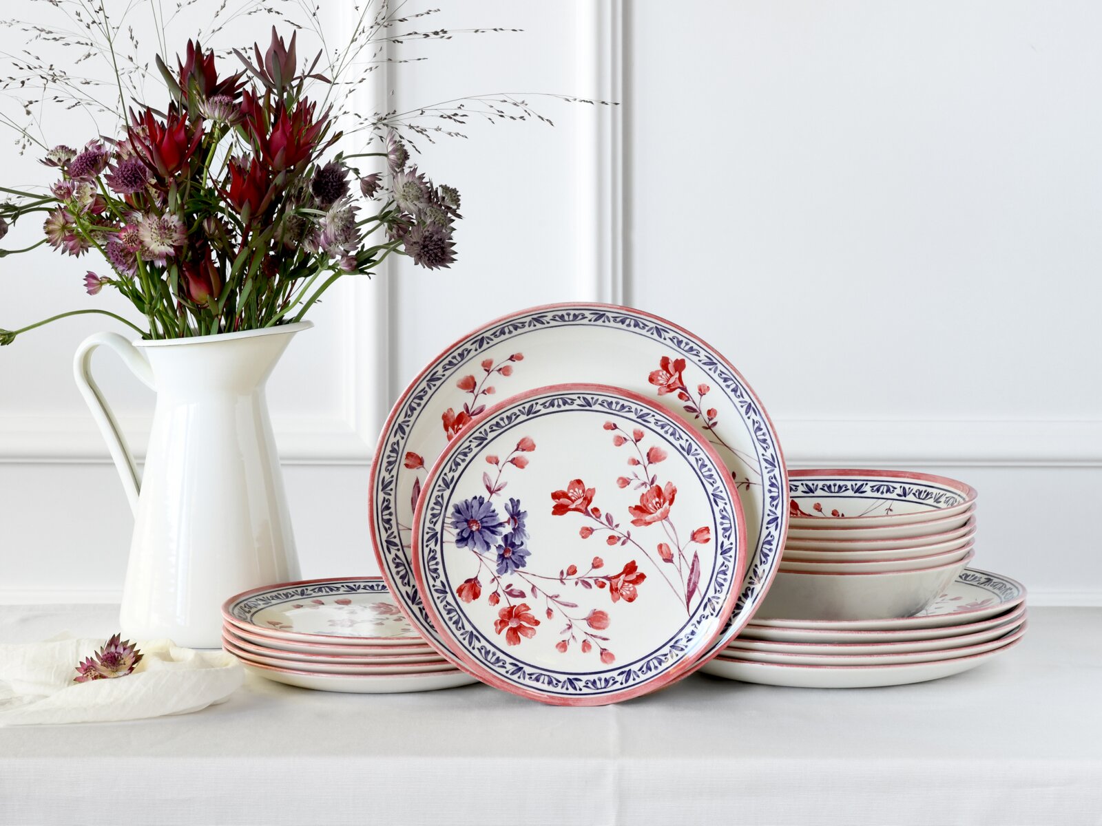 creaTable Tellerset 18-tlg COTTAGE Blumenporzellan-Geschirrset mit Tellern und Schalen in Weiß mit rotem und blauem Blütendekor, seitlich auf einem Tisch arrangiert, Frontansicht leicht schräg von vorn.