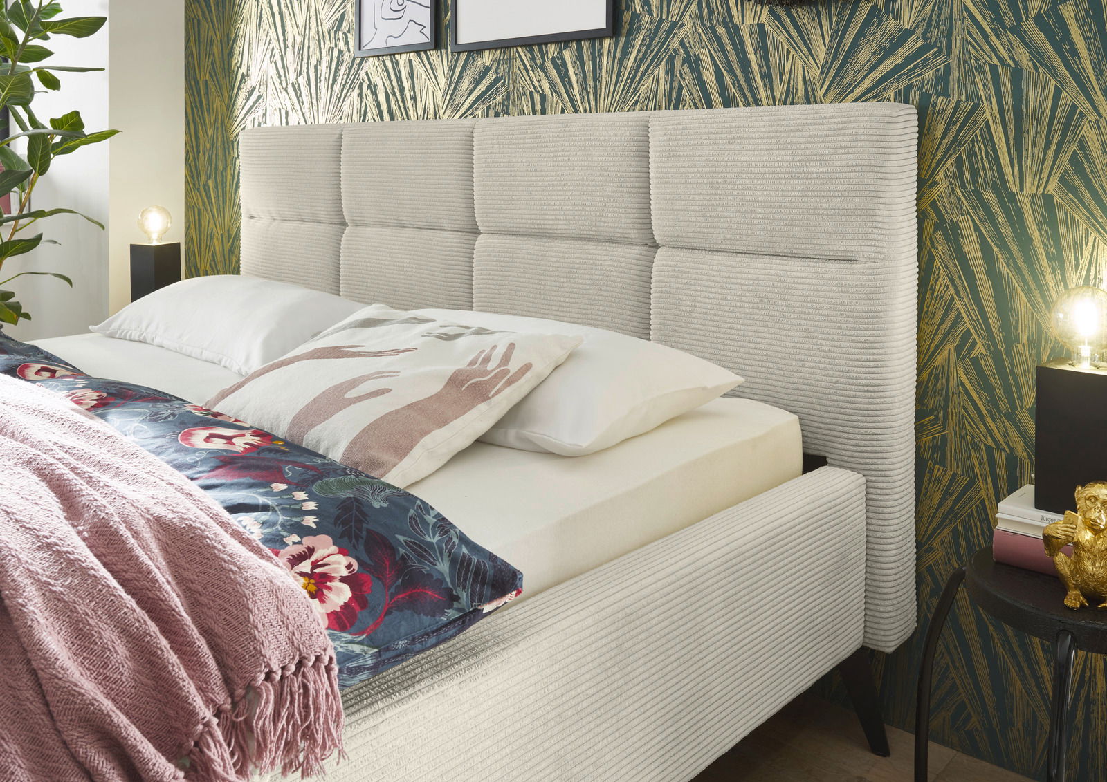 Seitliche Perspektive auf ein elegantes Polsterbett mit gestepptem Kopfteil, umgeben von stilvoller Dekoration und Pflanzen.