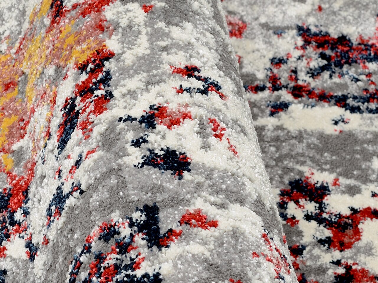 Nahaufnahme eines Designer-Teppichs mit abstraktem Muster in Grau, Rot, Gelb und Blau. Die Perspektive zeigt eine seitliche Ansicht der Textur und Farben.