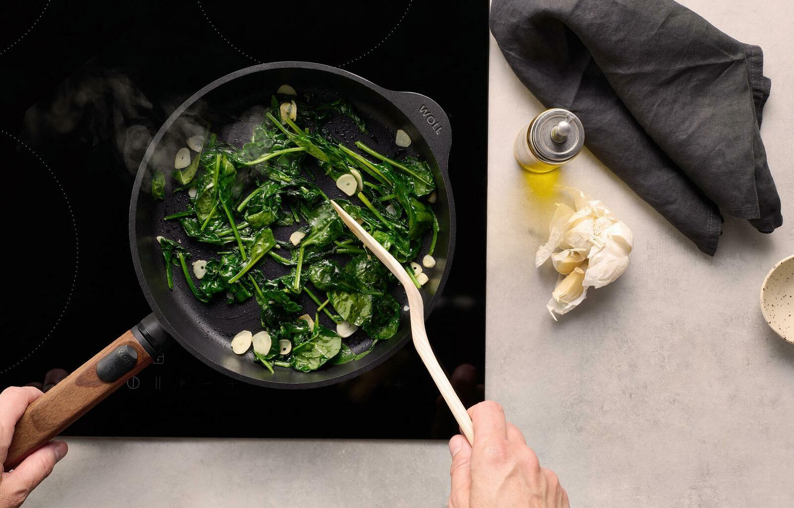 Gusseiserne Flachpfanne mit Holzgriff, von oben fotografiert, auf einem Herd mit Spinat und Knoblauch in der Pfanne.
