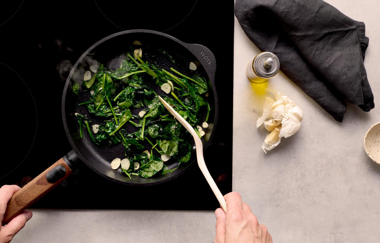 Woll Guss-Flachpfanne ECO LOGIC QXR Gusseiserne Flachpfanne mit Holzgriff, von oben fotografiert, auf einem Herd mit Spinat und Knoblauch in der Pfanne.