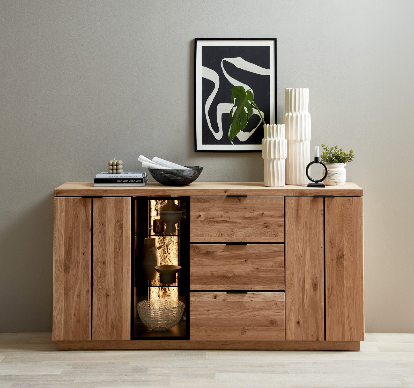 Cantus Sideboard DONNA Frontale Ansicht eines modernen Sideboards aus massivem Holz mit sichtbarer Maserung, zwei Türen, drei Schubladen und beleuchtetem Glasvitrinenfach; dekoriert mit Vasen, Kerzenhalter, Schale, Büchern und gerahmter abstrakter Wandkunst darüber.