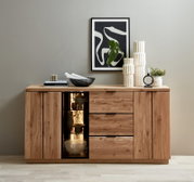 Frontale Ansicht eines modernen Sideboards aus massivem Holz mit sichtbarer Maserung, zwei Türen, drei Schubladen und beleuchtetem Glasvitrinenfach; dekoriert mit Vasen, Kerzenhalter, Schale, Büchern und gerahmter abstrakter Wandkunst darüber.