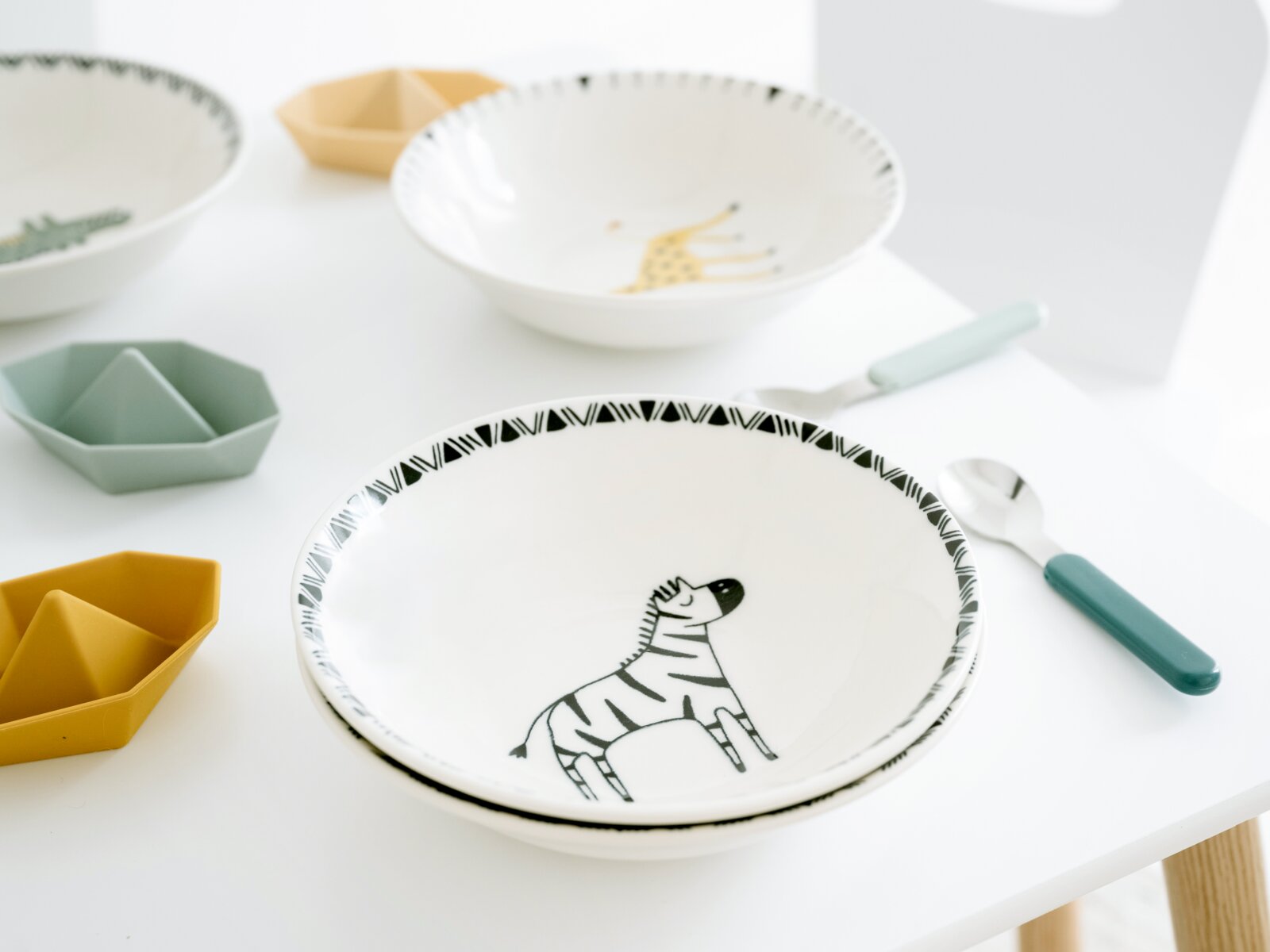 Weiße Kinderschale aus Keramik/Porzellan mit Zebra-Motiv und schwarzem Randmuster auf einem hellen Tisch, aufgenommen in leichter Draufsicht (schräg von oben) mit Besteck daneben