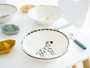 Weiße Kinderschale aus Keramik/Porzellan mit Zebra-Motiv und schwarzem Randmuster auf einem hellen Tisch, aufgenommen in leichter Draufsicht (schräg von oben) mit Besteck daneben
