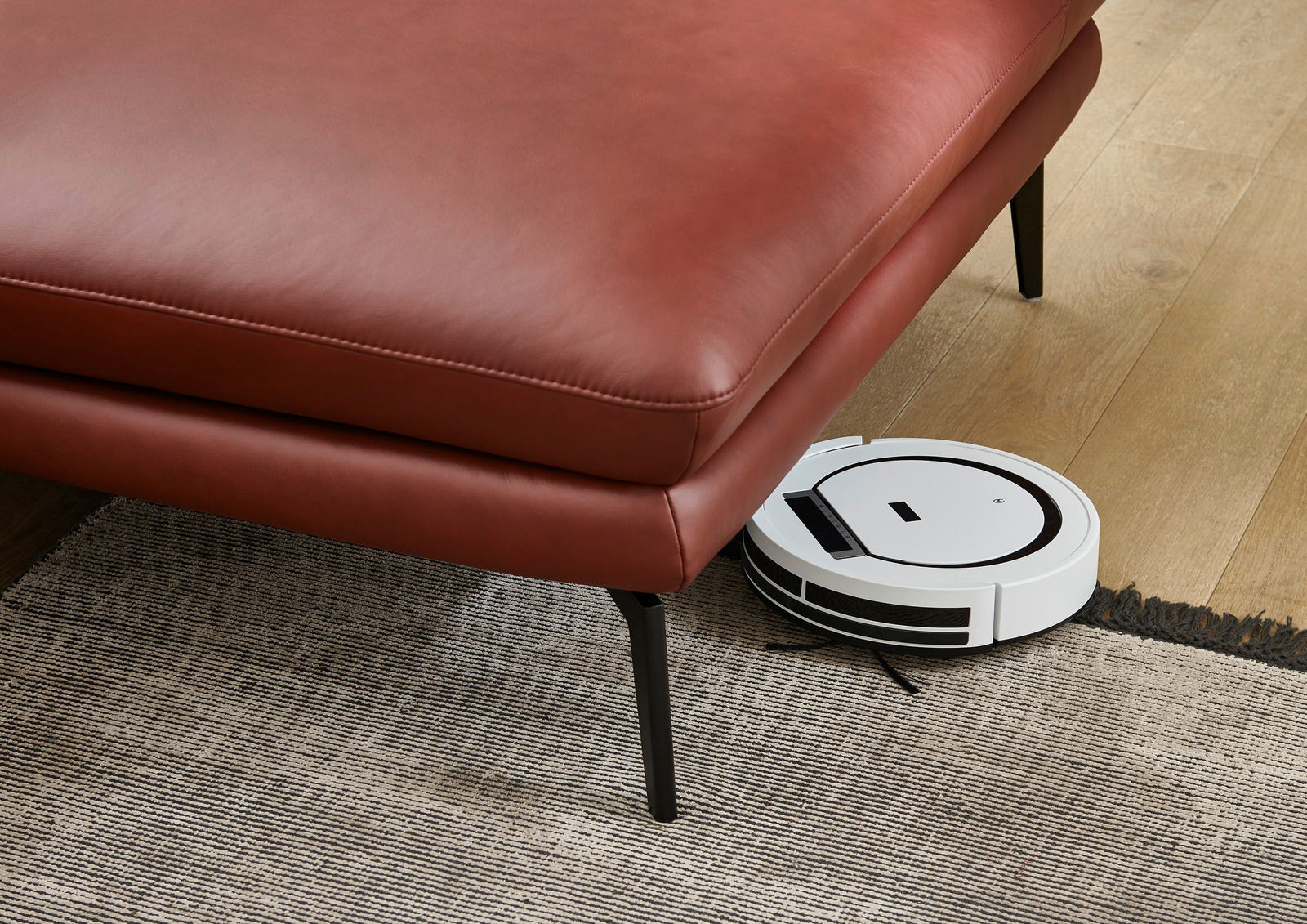 Rote Ledercouch mit schwarzen Beinen, darunter ein weißer Saugroboter auf einem Holzboden. Perspektive von schräg oben.