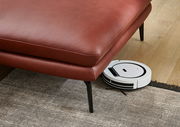 Rote Ledercouch mit schwarzen Beinen, darunter ein weißer Saugroboter auf einem Holzboden. Perspektive von schräg oben.