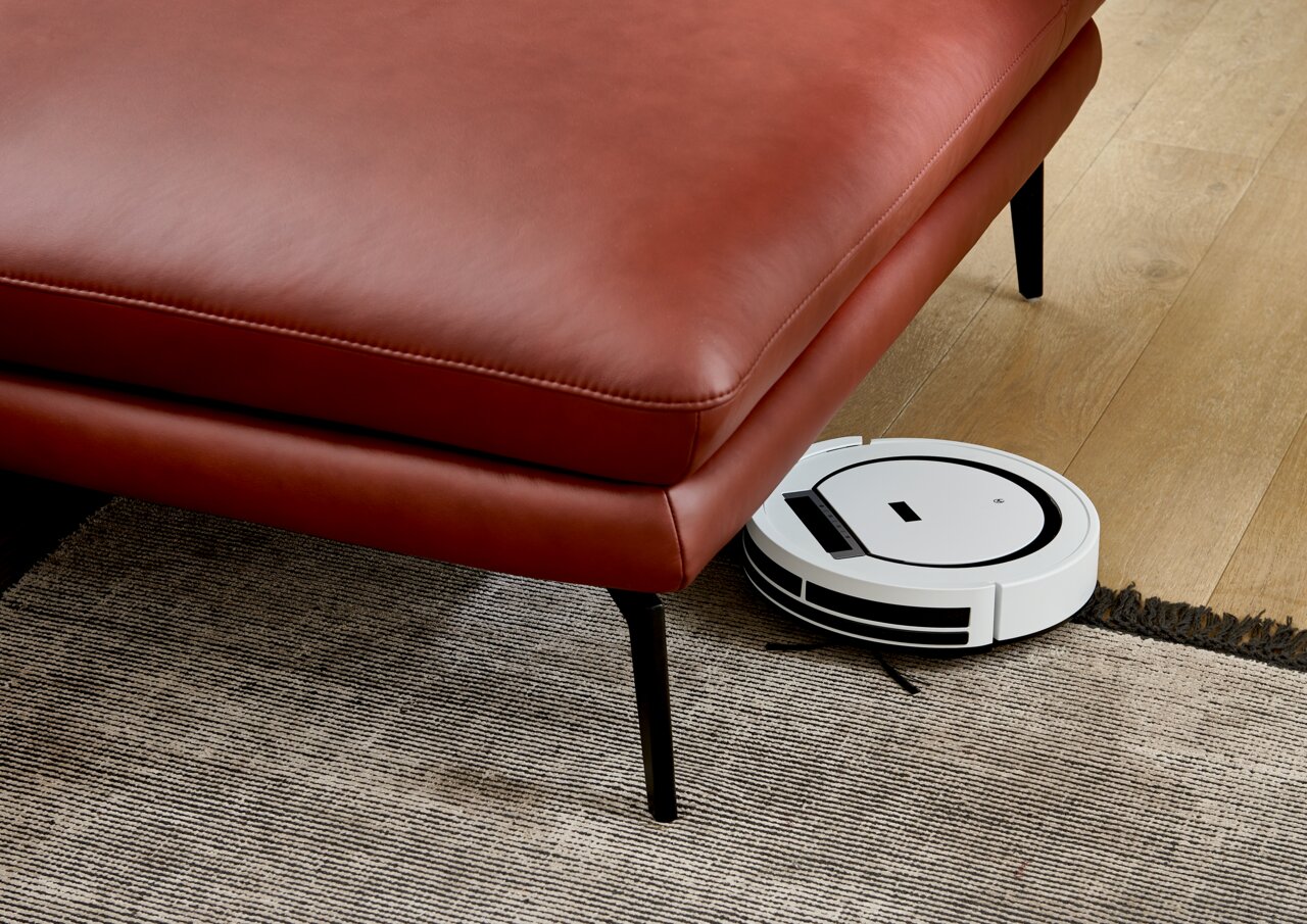 Rote Ledercouch mit schwarzen Beinen, darunter ein weißer Saugroboter auf einem Holzboden. Perspektive von schräg oben.