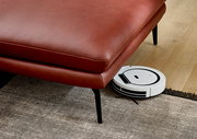 Rote Ledercouch mit schwarzen Beinen, darunter ein weißer Saugroboter auf einem Holzboden. Perspektive von schräg oben.