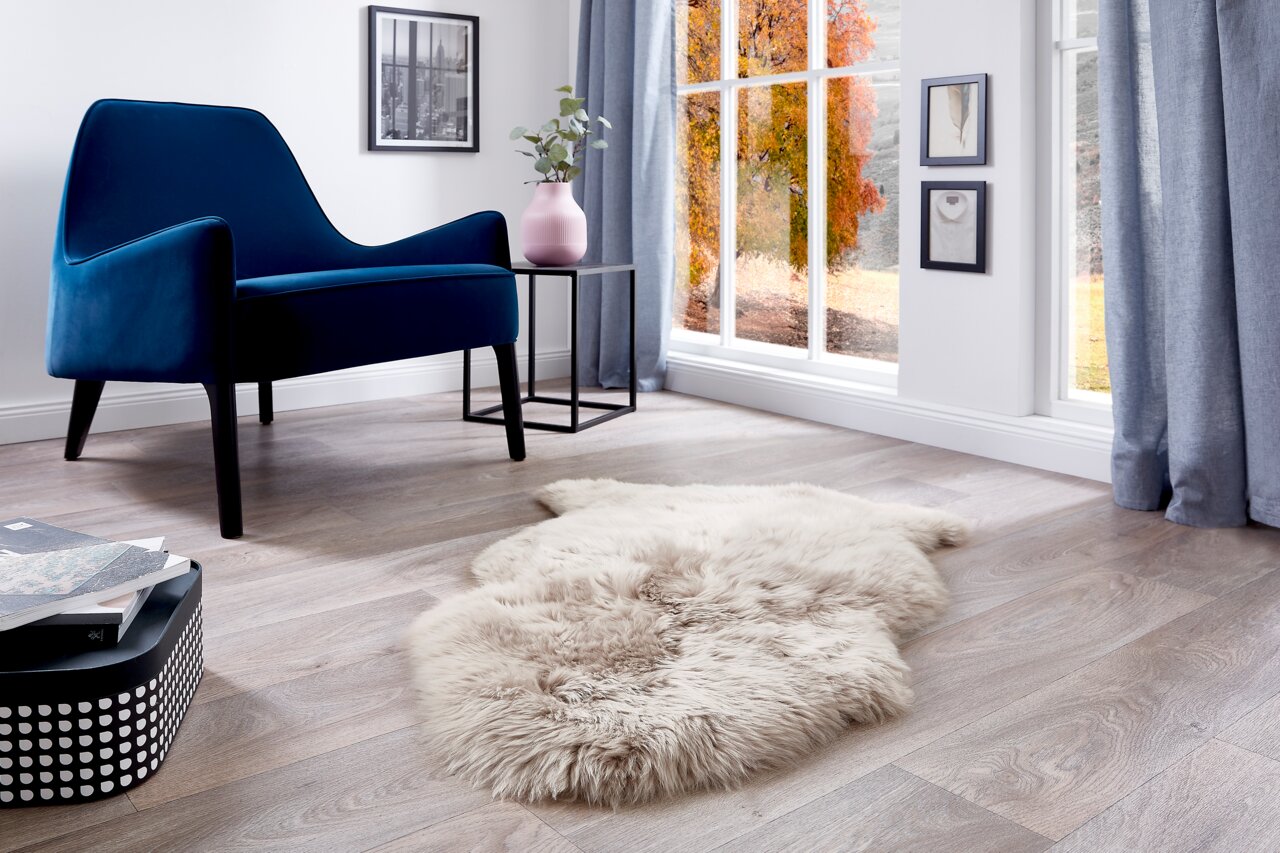 Gemütliches Schaffell auf einem Holzboden in einem stilvoll eingerichteten Wohnzimmer mit einem blauen Sessel und großen Fenstern, seitliche Perspektive.